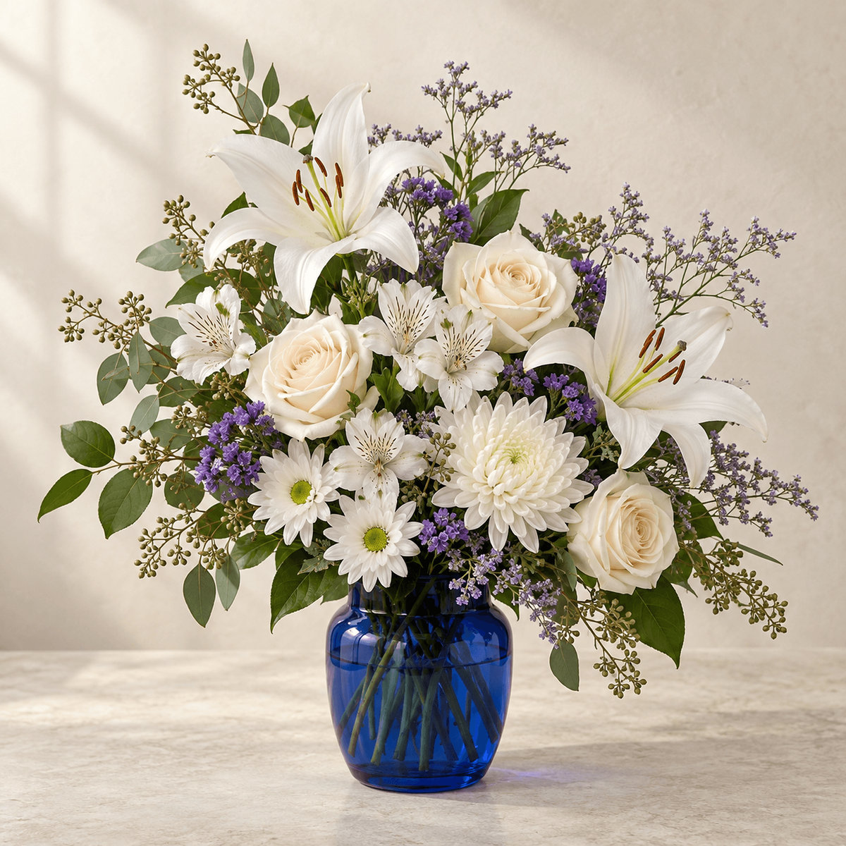 White lilies, cream roses, white chrysanthemums and alstroemeria with violet accents in a rounded cobalt-blue glass vase.