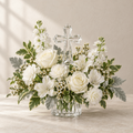 A clear crystal cross rises above white and cream flowers with fresh green and silver foliage. A thoughtful expression of faith for the family home.