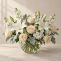 White lilies, cream roses, white stock and soft silver-green foliage in a round clear glass vase.