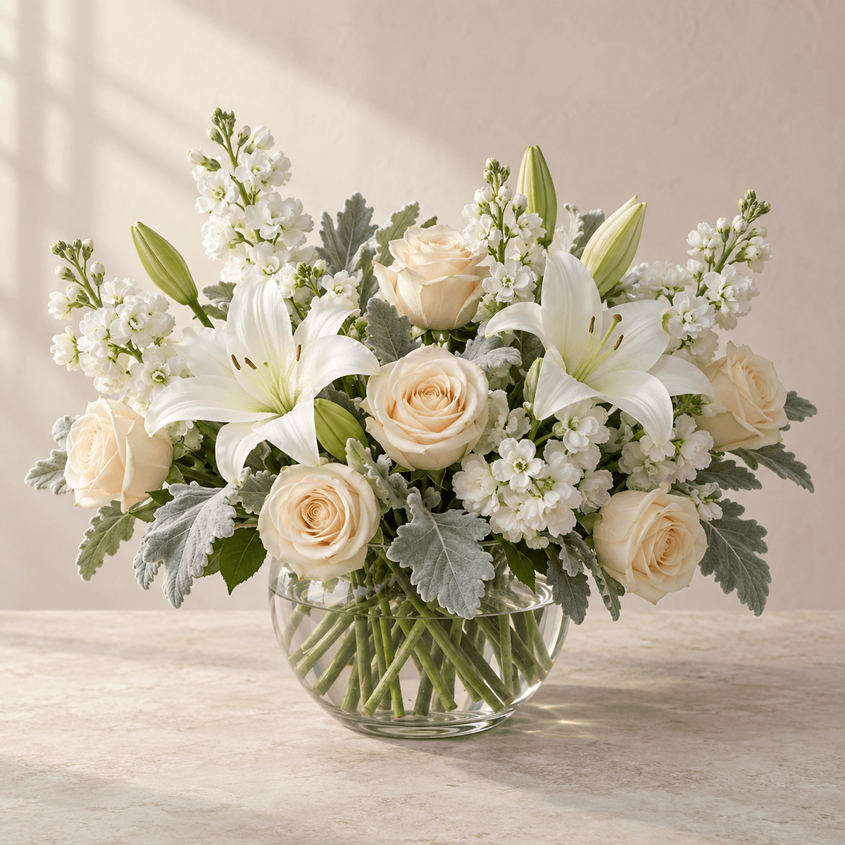 White lilies, cream roses, white stock and soft silver-green foliage in a round clear glass vase.